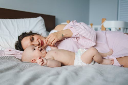 Woman in Pink Dress Lying on Bed Next to Baby in Diapers